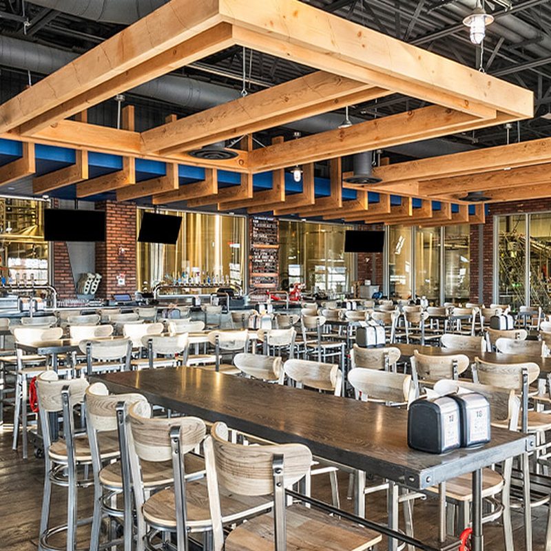 Open brewery dining hall with wood beams, communal tables, and brewery tanks visible through the back wall