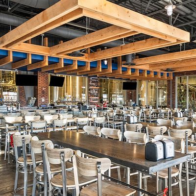 Open brewery dining hall with wood beams, communal tables, and brewery tanks visible through the back wall