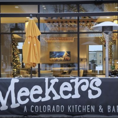 Evening view through the storefront windows into the warm-lit dining room with string lights and a Colorado mountain mural