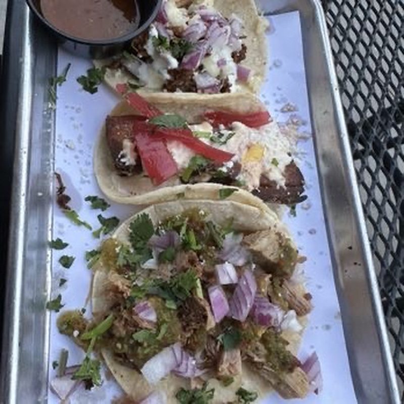Three tacos on a metal tray with different fillings, salsas, and cilantro garnish.